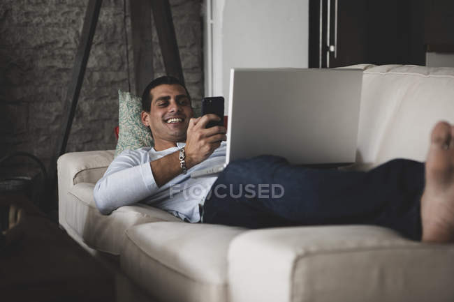 Счастливый молодой человек лежит дома на диване с помощью мобильного телефона и ноутбука — стоковое фото