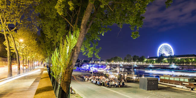 Francia, París, Campos Elíseos, Quai Anatole, gente en la orilla del río Sena por la noche - foto de stock