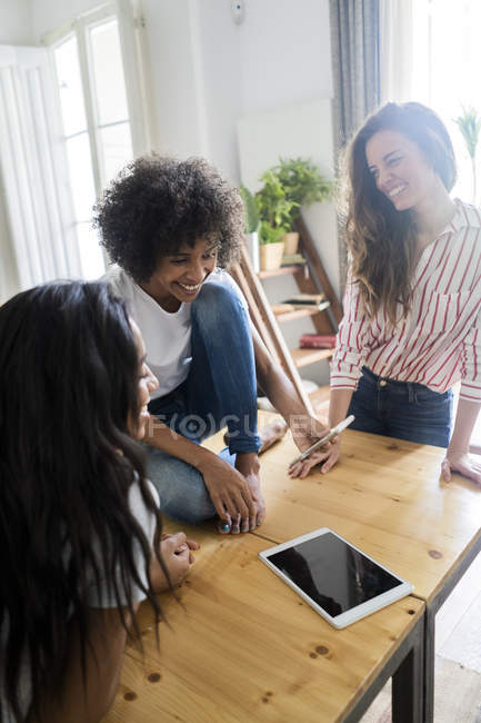Tres mujeres felices mirando el teléfono celular en la mesa en casa - foto de stock