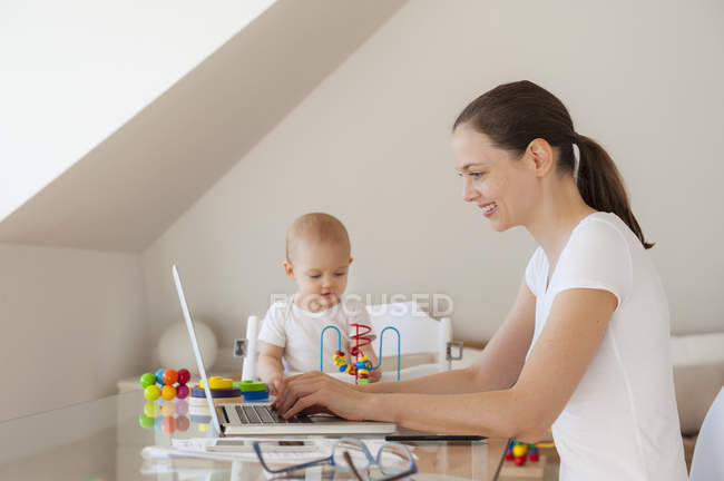 Lächelnde Mutter mit Laptop und kleine Tochter spielen zu Hause am Tisch — Stockfoto
