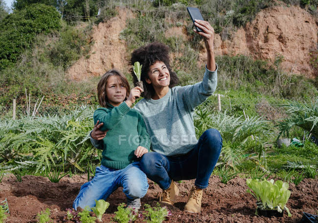 Mãe tomando selfies de plantio de mudas de alface em uma horta, com seu filho — Fotografia de Stock