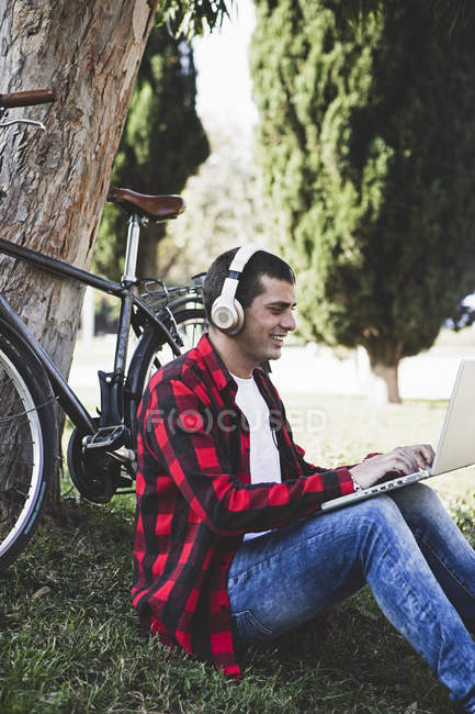 Young man sitting in a park wearing headphones and using laptop next to bicycle — Stock Photo