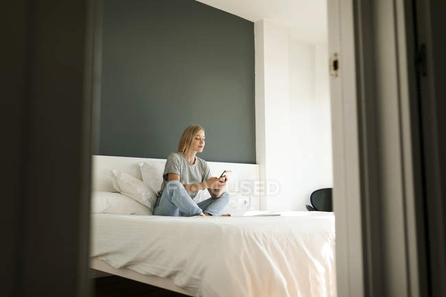 Jeune femme assise sur le lit en utilisant un téléphone portable — Photo de stock