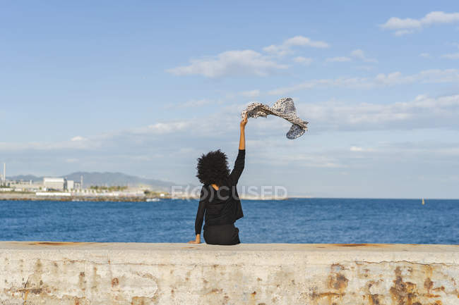 Spain, Barcelona, back view of woman dressed in black sitting on wall waving with scarf — Stock Photo