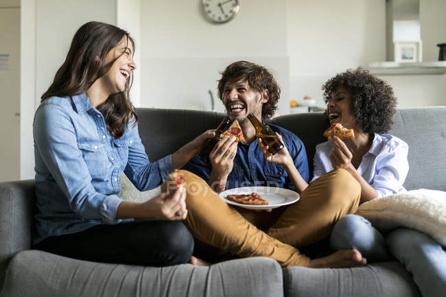 Веселые друзья сидят на диване, пьют пиво и едят пиццу — стоковое фото