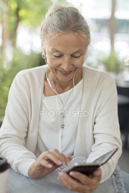 Mulher sênior sorridente em um café ao ar livre com telefone celular e fones de ouvido — Fotografia de Stock