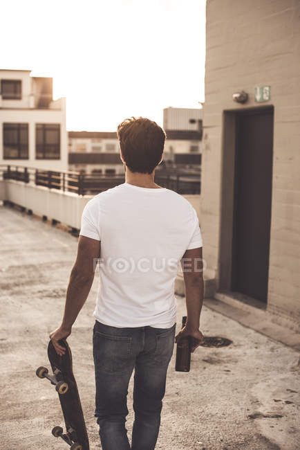 Back view of young man with skateboard and beer bottle on roof terrace at evening twilight — Stock Photo