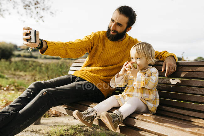 Père et fille assis sur un banc dans le parc en automne, père prenant un selfie, fille mangeant une pomme — Photo de stock