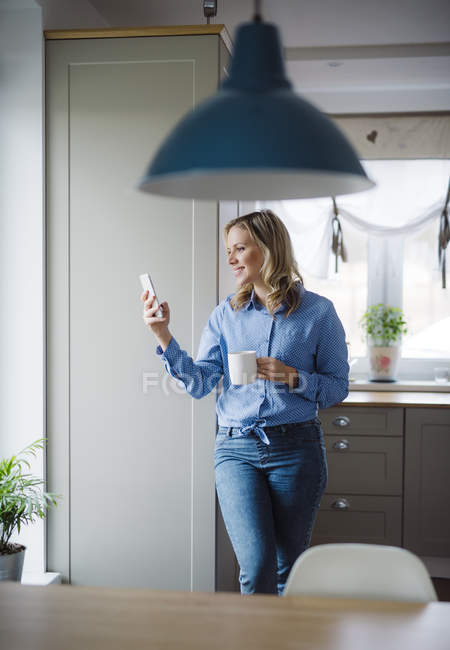 Улыбающаяся женщина держит дома чашку кофе и смартфон — стоковое фото