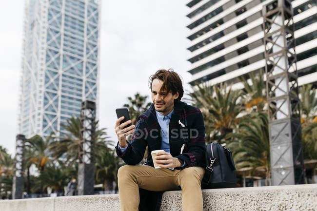 Spain, Barcelona, smiling man sitting in the city with takeaway coffee and cell phone — Stock Photo