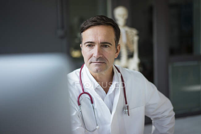 Arzt in der Arztpraxis betrachtet Computer mit Skelett im Hintergrund — Stockfoto