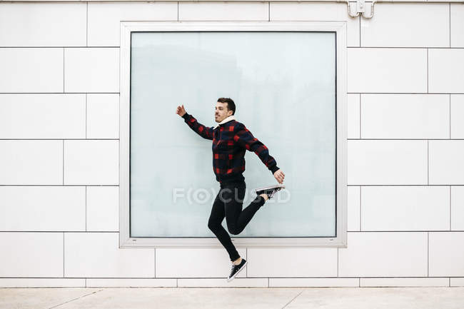 Junger Mann in lässiger Kleidung springt mit einer weißen Wand und einem großen Fenster im Hintergrund — Stockfoto