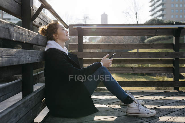 Jeune femme se reposant à l'extérieur tenant un téléphone cellulaire — Photo de stock