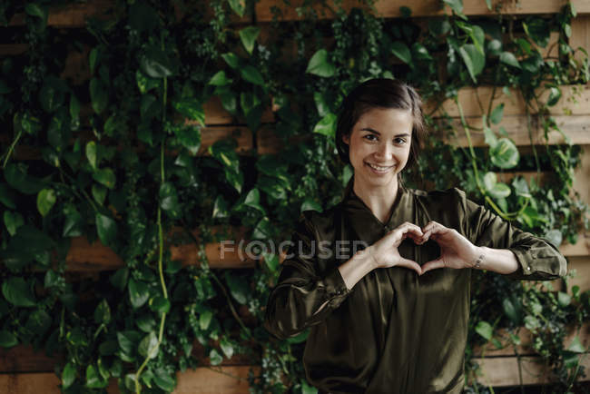 Portrait de jeune femme souriante façonnant le cœur au mur avec des plantes grimpantes — Photo de stock