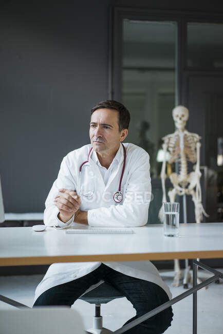 Medico seduto alla scrivania nella pratica medica con scheletro in background — Foto stock