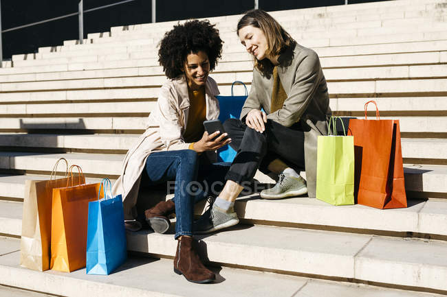 Due donne felici con le borse della spesa seduti sulle scale utilizzando il telefono cellulare — Foto stock