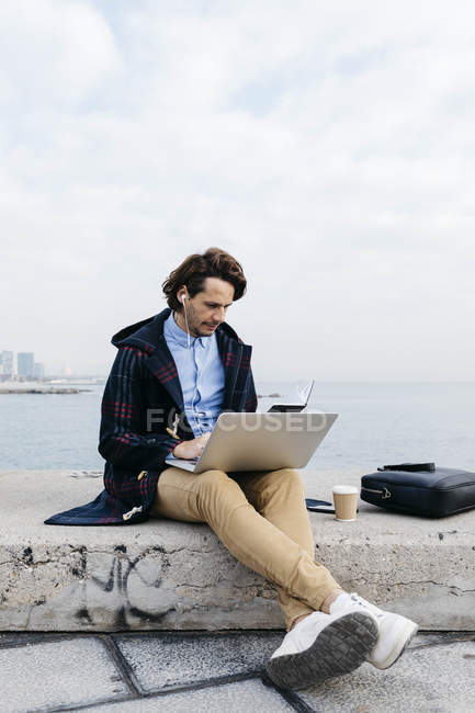 Іспанія, Барселона, людина, що сидить у морі, працює з ноутбуком і зошитом. — стокове фото