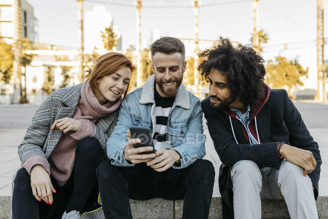 Троє щасливих друзів сидять надворі і дивляться на мобільний телефон. — стокове фото