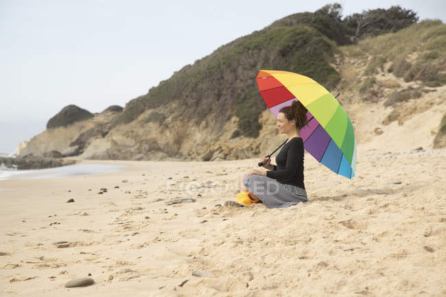 Mujer con paraguas colorido sentada en la playa - foto de stock