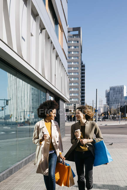 Two happy women with shopping bags and takeaway coffee walking in the city — Stock Photo