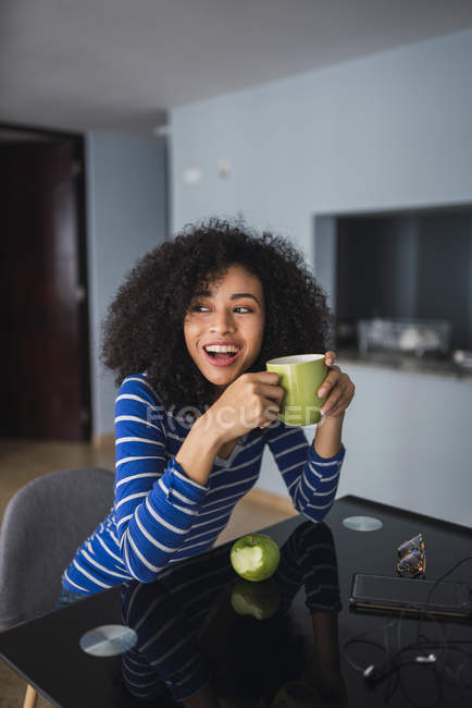 Portrait of laughing young woman drinking coffee and eating an apple at home — Stock Photo