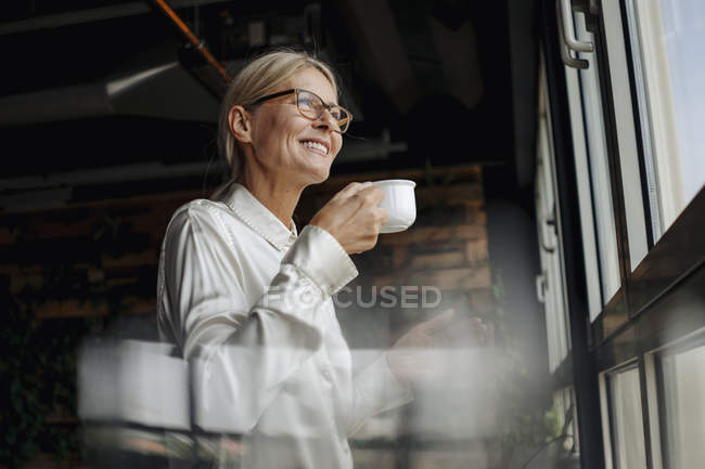 Souriante femme d'affaires tenant tasse de café regardant par la fenêtre — Photo de stock