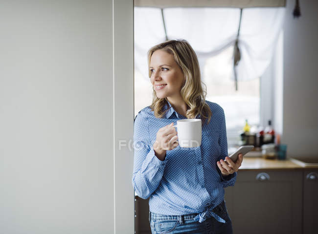 Mujer sonriente sosteniendo una taza de café y teléfono inteligente en casa - foto de stock