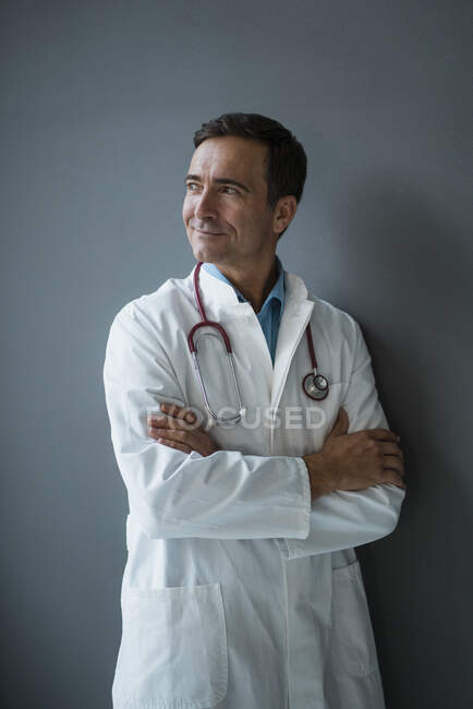 Médico sorridente em pé em uma parede cinza olhando para os lados — Fotografia de Stock