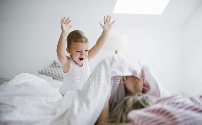 Feliz hijo pequeño jugando con la madre en la cama en casa - foto de stock