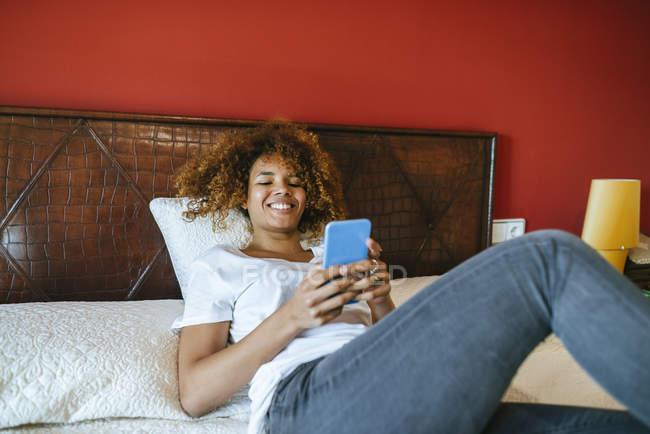 Felice giovane donna con i capelli ricci sdraiata a letto a casa utilizzando il telefono cellulare — Foto stock