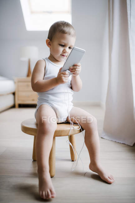 Малыш сидит дома на стуле, используя смартфон. — стоковое фото