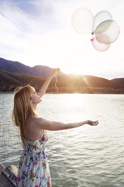 Young woman wearing summer dress with floral design standing on jetty looking at balloons — Stock Photo