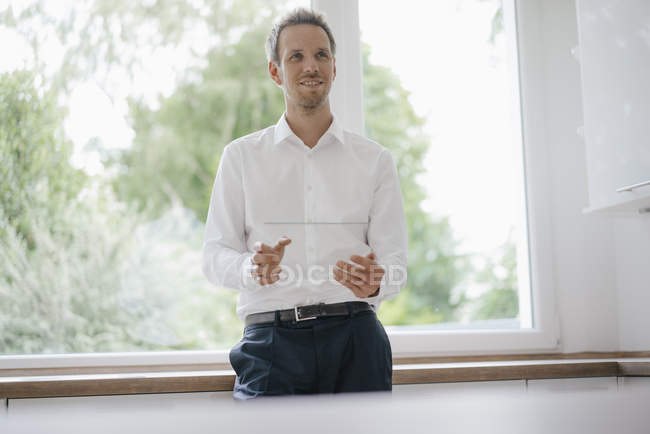 Бізнесмен стоїть біля кухонного вікна, за допомогою скляного сенсорного екрану, посміхається — стокове фото