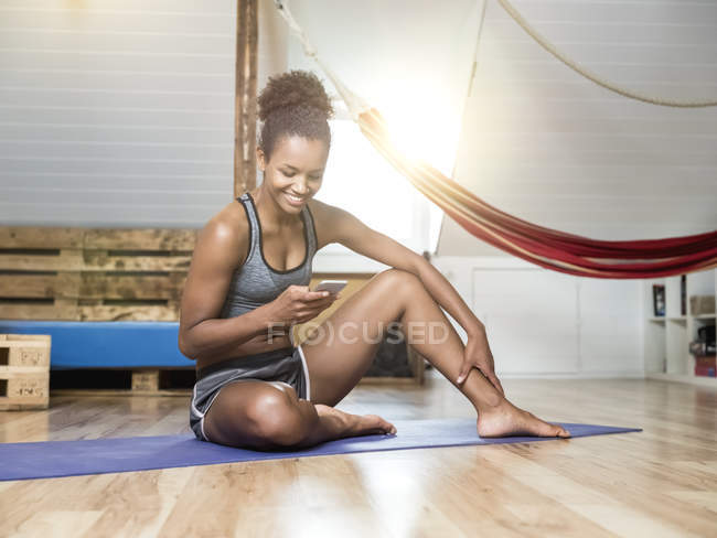 Smiling young woman sitting on yoga mat looking at cell phone — Stock Photo