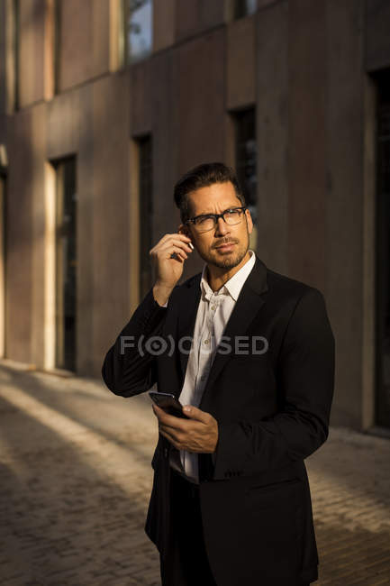 Hombre de negocios con teléfono celular y auriculares en la ciudad mirando a su alrededor - foto de stock