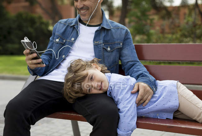 Portrait d'un garçon souriant avec son père utilisant un téléphone portable et des écouteurs sur un banc — Photo de stock