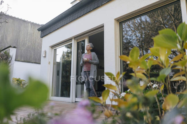 Mujer madura con taza de café de pie en la puerta de la terraza - foto de stock