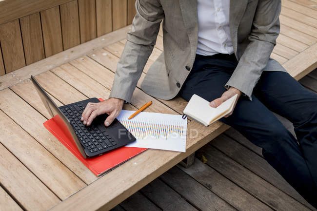 Крупный план бизнесмена, сидящего на скамейке с помощью ноутбука и дневника — стоковое фото
