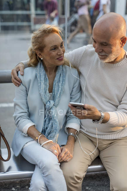 Spain, Barcelona, senior couple sitting at tram stop in the city sharing smartphone with earbuds — Stock Photo