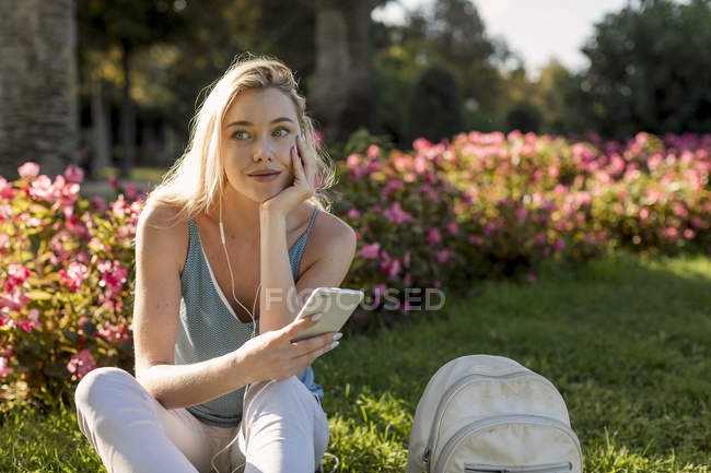 Jeune femme souriante assise dans le parc avec téléphone cellulaire et écouteurs — Photo de stock