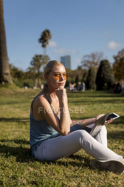 Молода жінка сидить в парку з мобільним телефоном і ноутбуком. — стокове фото