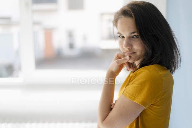 Mulher bonita de pé contra a janela, com a mão no queixo, pensando — Fotografia de Stock
