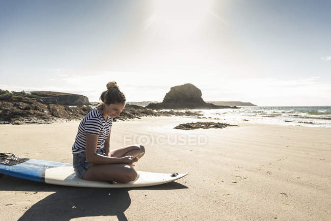 Young woman on the beach, sitting on surfboard, using smartphone — Stock Photo