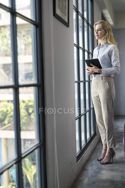 Довірлива ділова жінка з планшетом стоїть в офісі дивлячись у вікно — стокове фото