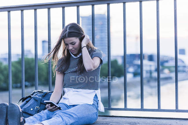 Mujer joven sentada en un puente mirando el teléfono celular - foto de stock