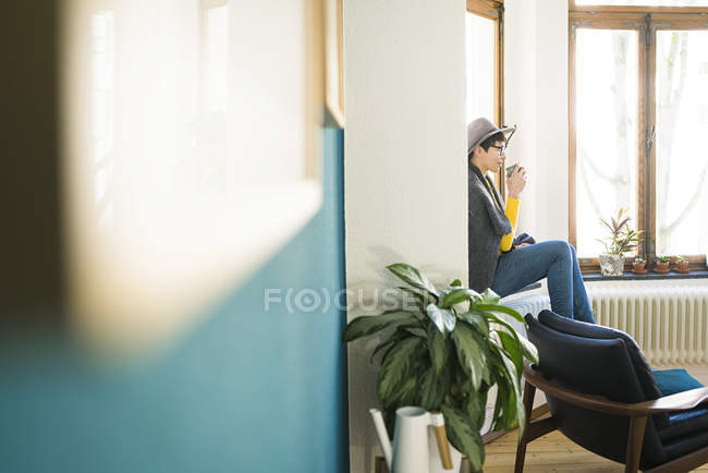 Frau sitzt auf Fensterbank in stilvoller Wohnung und hält Kaffeebecher — Stockfoto