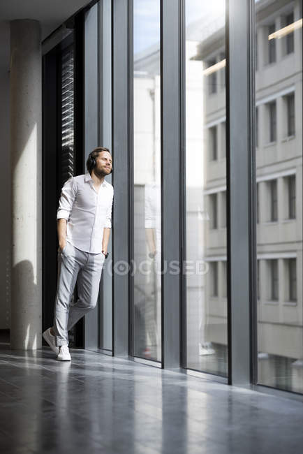 Homem de negócios casual inclinado para a janela no espaço de escritório moderno com os olhos fechados ouvir podcast — Fotografia de Stock