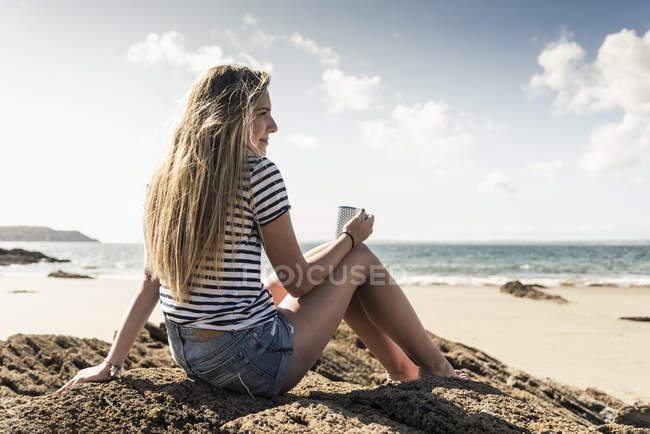 Junge Frau sitzt auf einem Felsen am Strand und entspannt sich bei einer Tasse Tee — Stockfoto