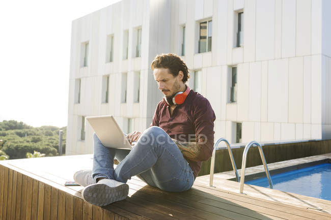 Homem sentado de pernas cruzadas em um terraço no último piso, usando laptop, usando fones de ouvido — Fotografia de Stock