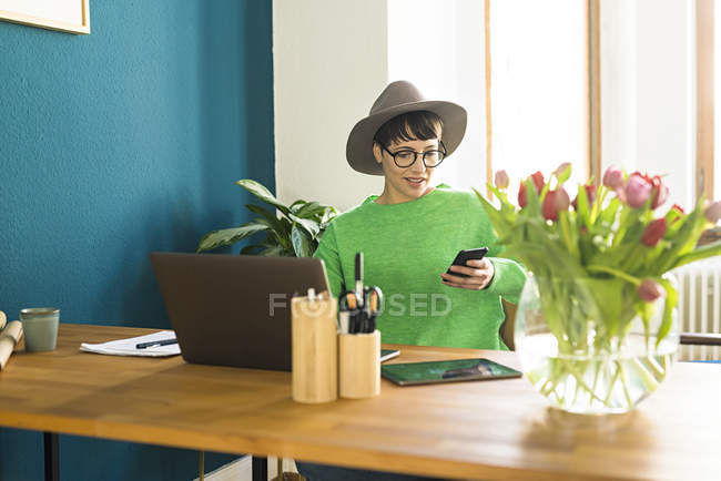 Businesswoman in home office working at her laptop and looking on her phone — Stock Photo